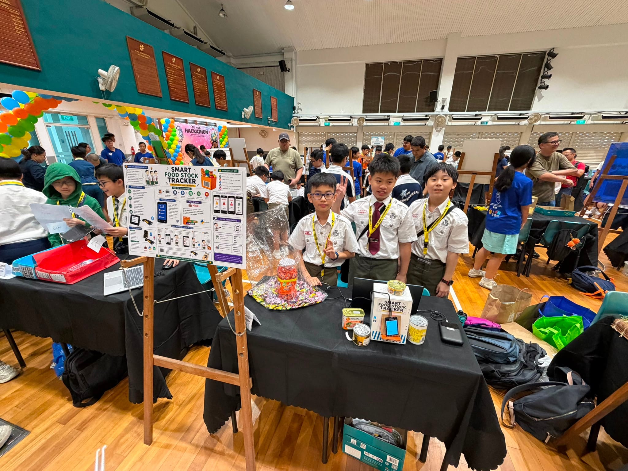 The photo shows pupils presenting a “Smart Food Stock Tracker” project at a busy school hackathon. It highlights teamwork, innovation, problem-solving, and applying technology to real-world needs.